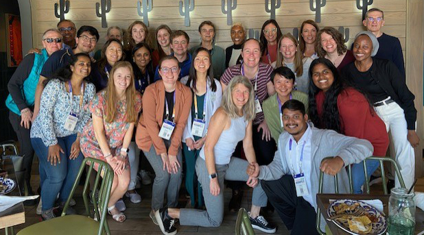 Large group of alumni and current lab members smiling and posing together indoors, many wearing conference name badges.